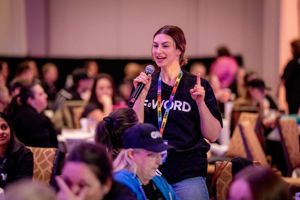 Member stands while talking into a mic and pointing upwards.