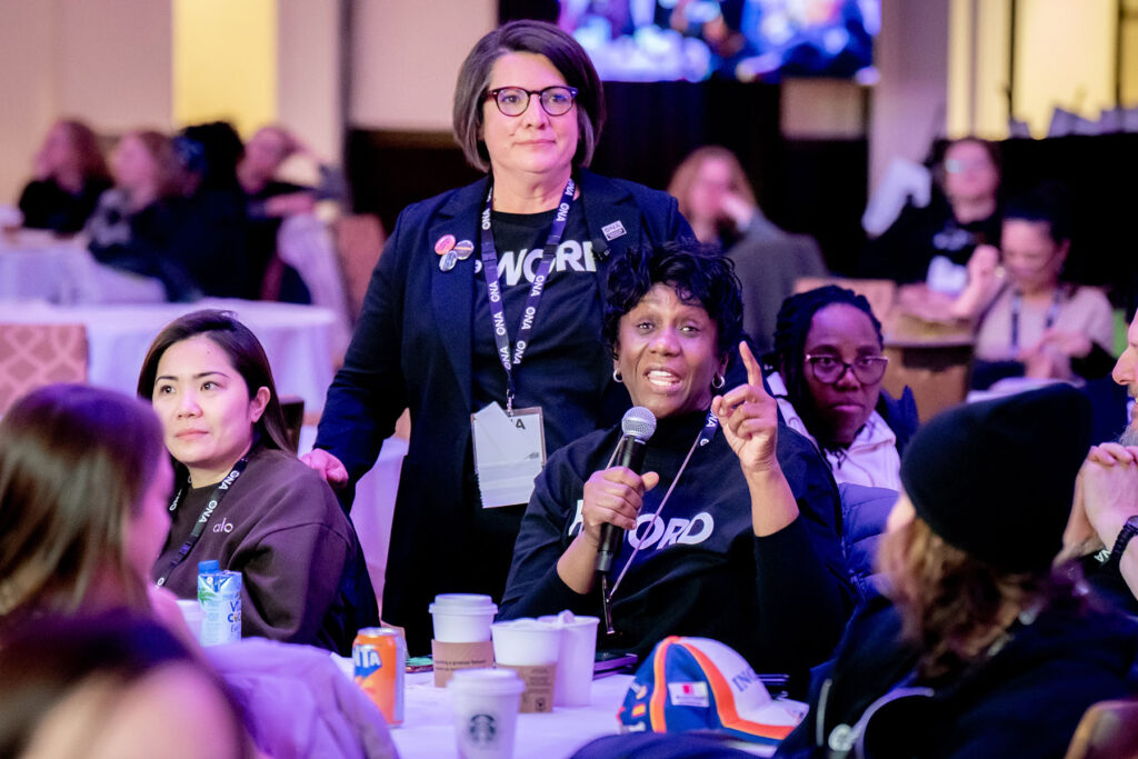 ONA Provincial President Erin Ariss stands beside a member sitting at a table and talking into a mic.