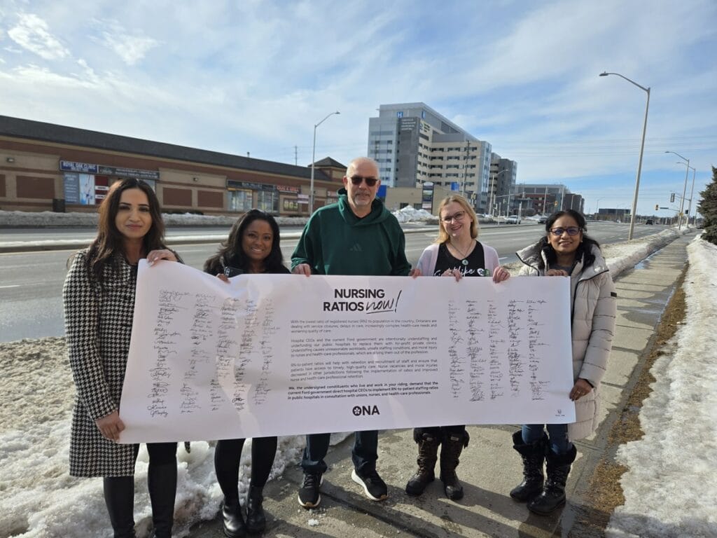 Members and candidate stand on sidewalk holding life-sized banner full of signatures.