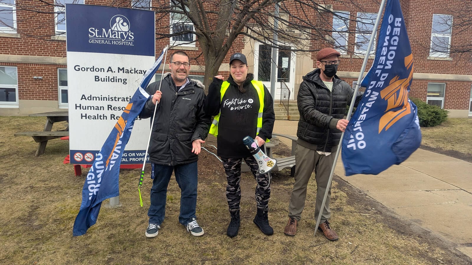 St. Mary’s General Hospital Bargaining Unit President Stephanie Hamill gives a fist pump as two men hold a life-sized banner full of signatures.