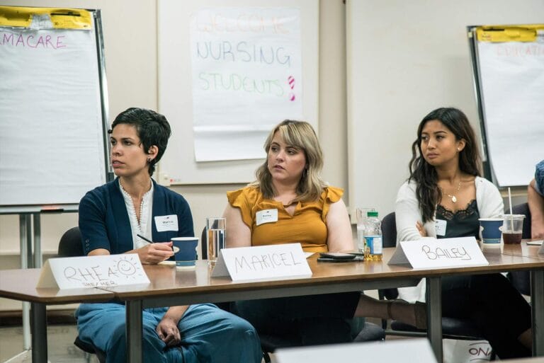 Three nursing students seated at a table look to front of room.