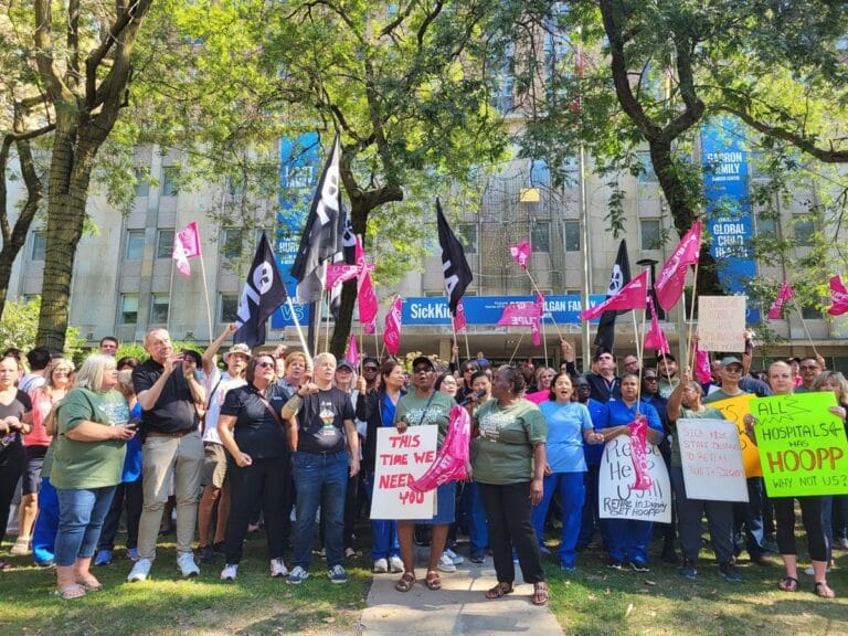 Large group of members, union allies and Sickkids health-care workers hold flags and pose for camera outside of hospital.
