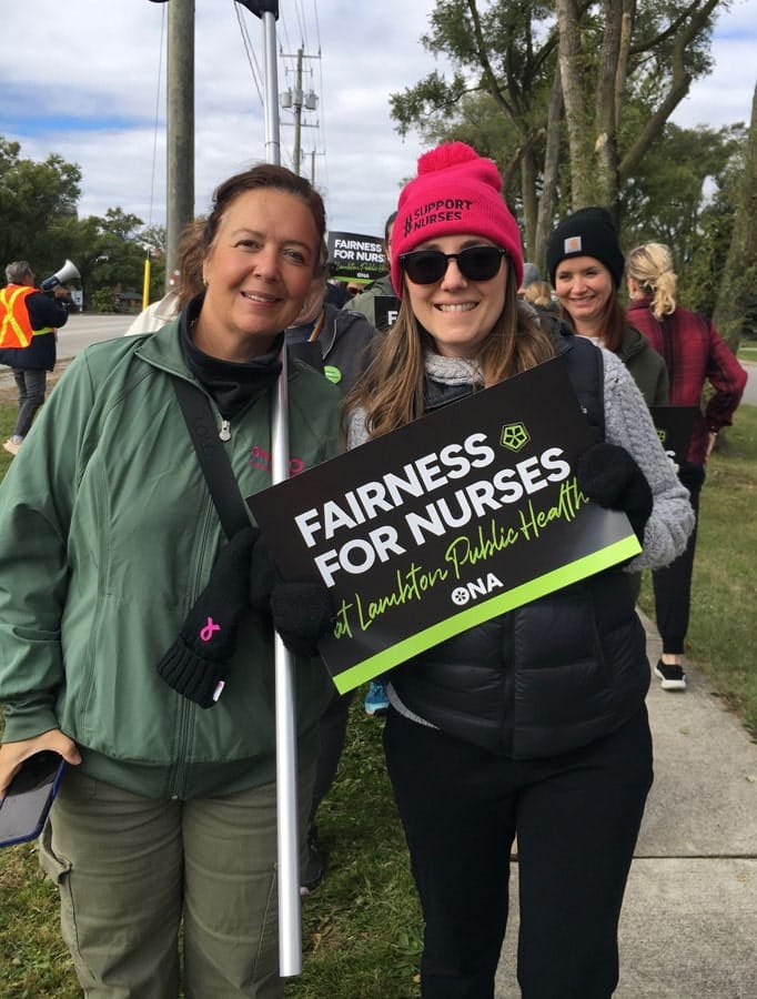 Two smiling members stop on the sidewalk to pose for camera, one carrying a sign.