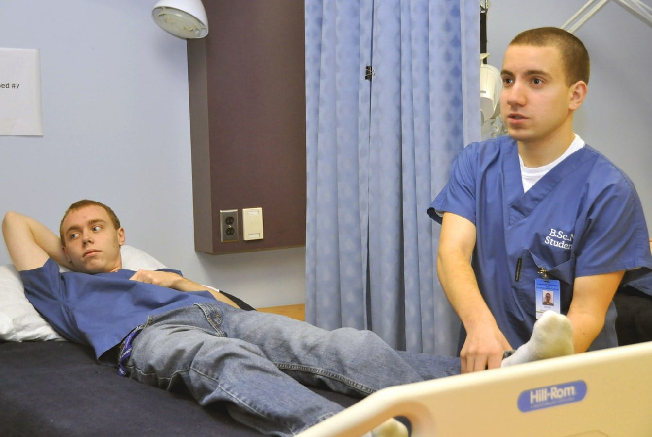 Nursing student lays in mock hospital bed while another stands beside him.