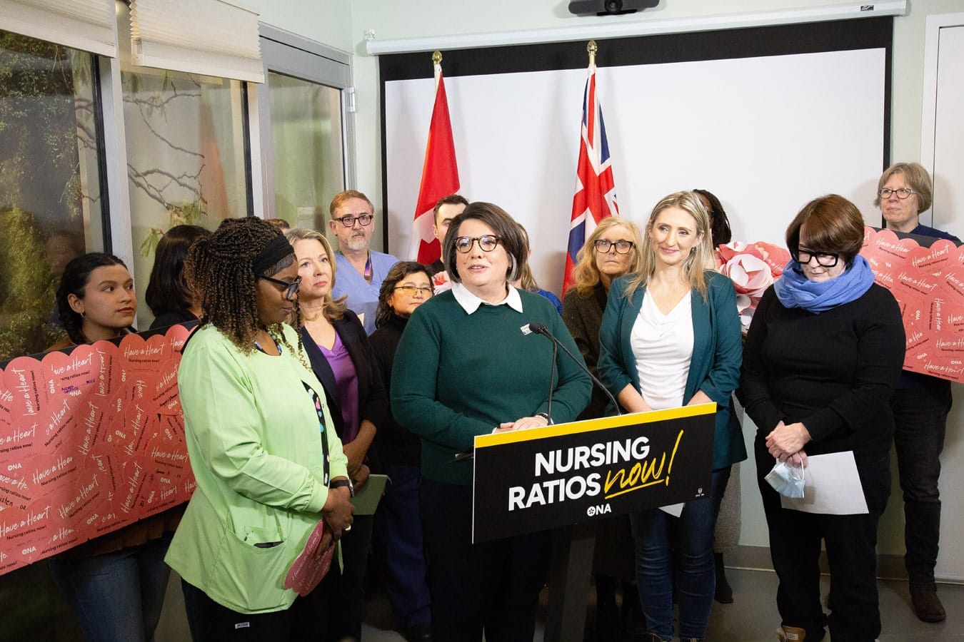 ONA Provincial President Erin Ariss at podium of media conference is surrounded by members holding valentines, Ontario NDP Leader Marit Stiles and Ontario Federation of Labour President Laura Walton.