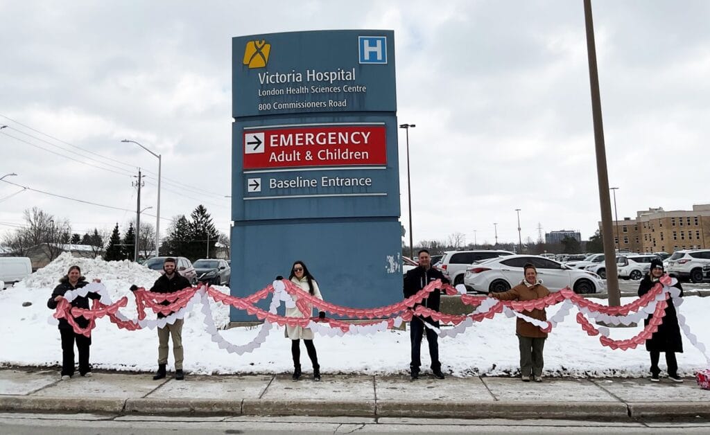 Members stand in a row under hospital outdoor sign holding a long string of valentines.