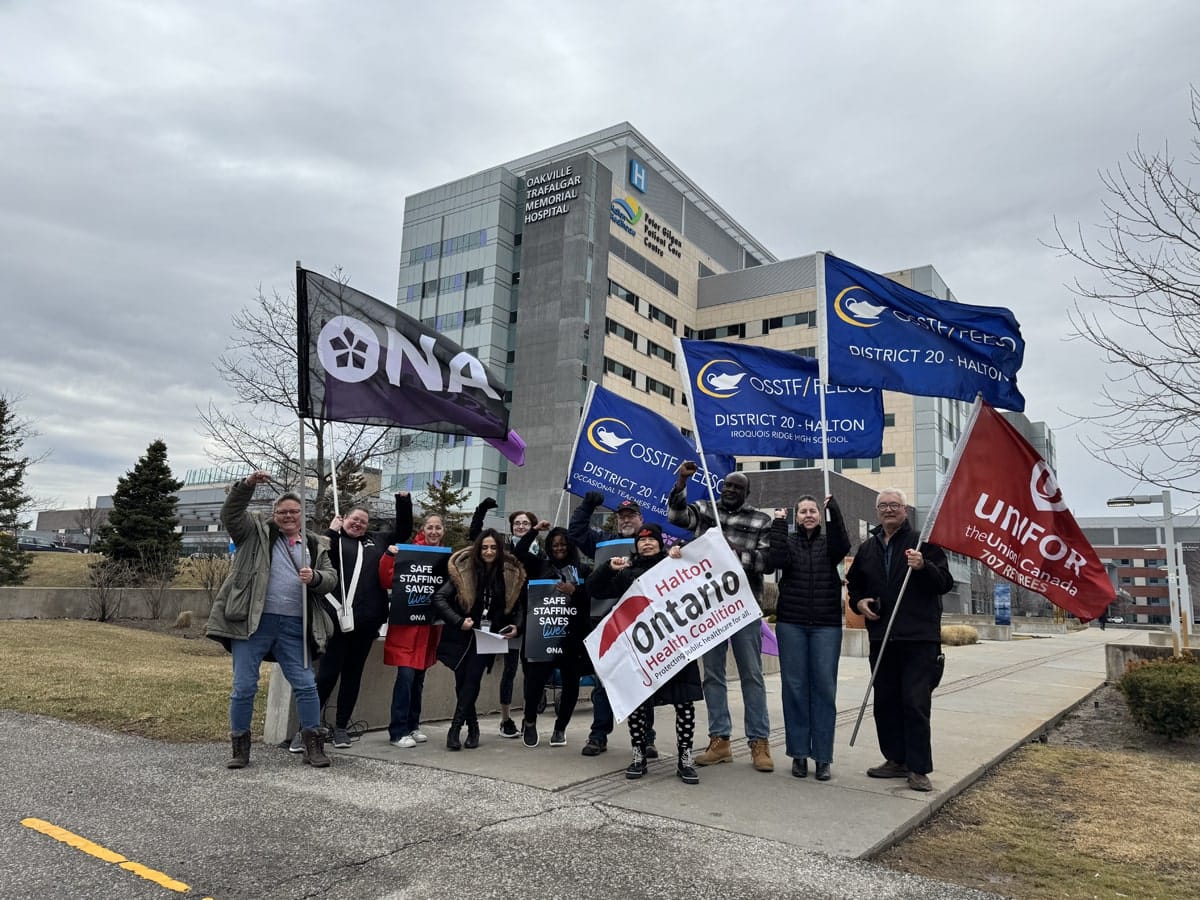 Group of members, staff and allies gather holding signs with hospital in background.
