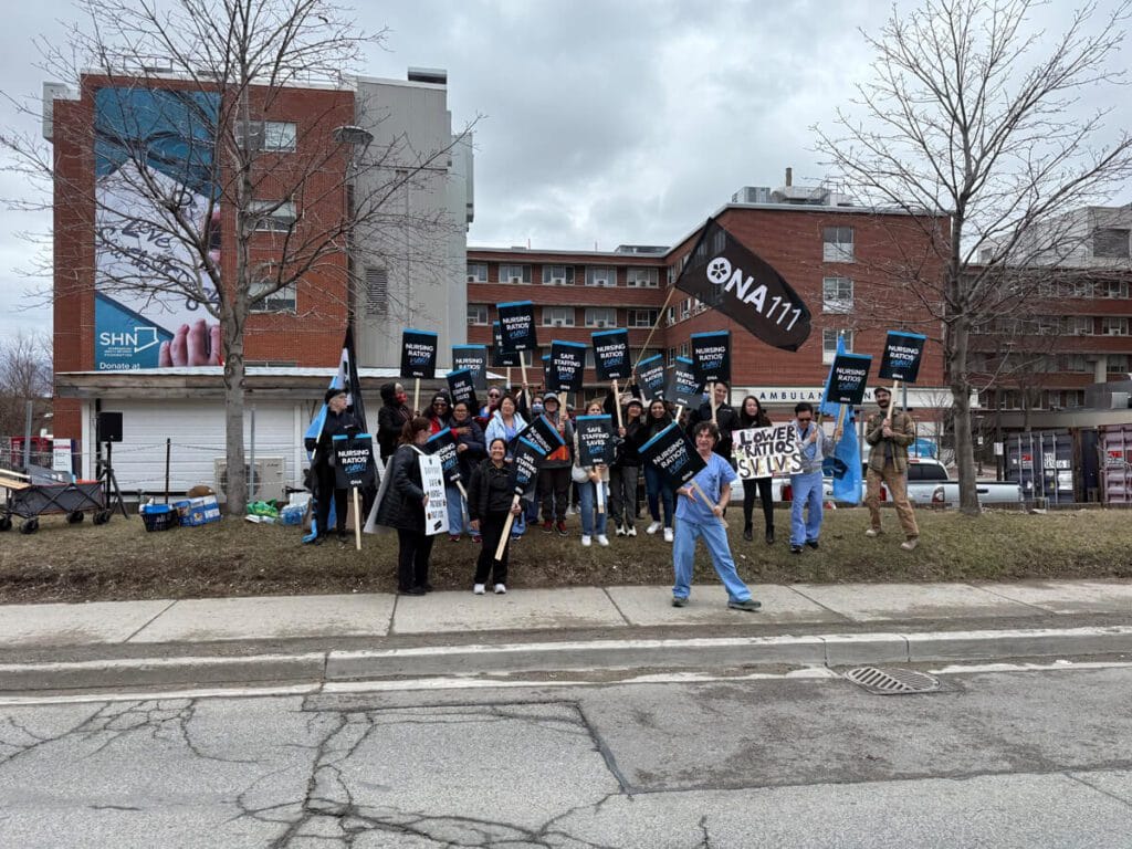 Group of members wearing scrubs and coats carry signs and union flags.