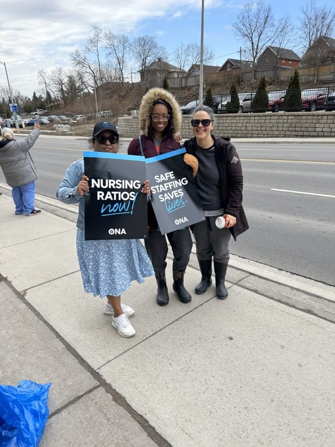 Three members stand together on sidewalk holding sign.