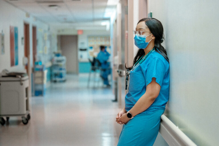 A female nurse wearing a mask leans against the railing in a hospital hallway.