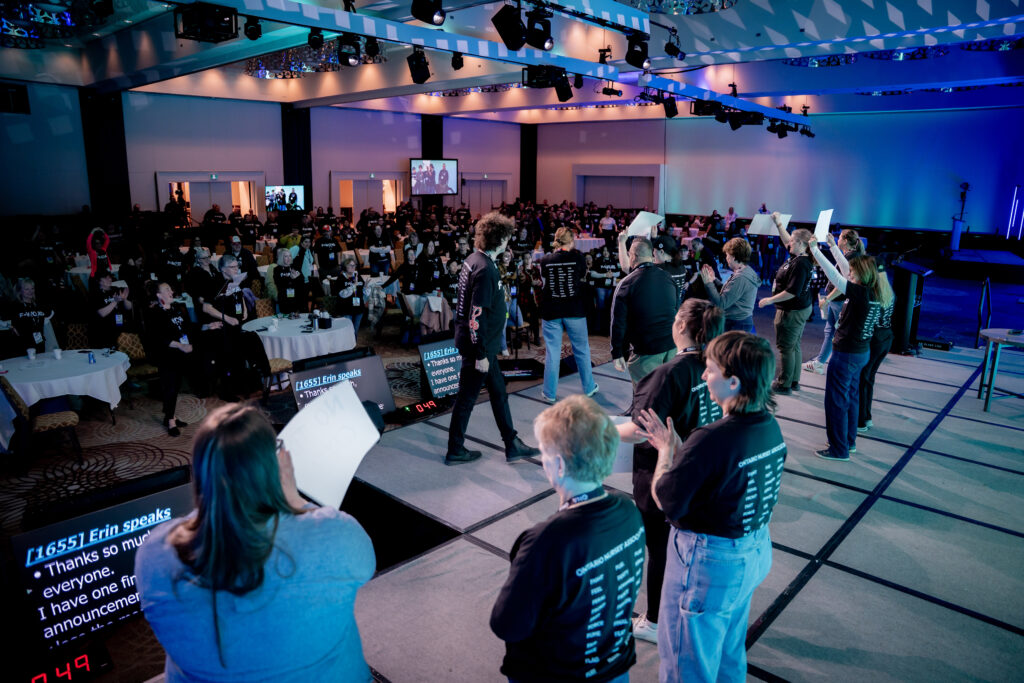 Thirteen people standing on a wide stage with the crowd they are addressing in the background.