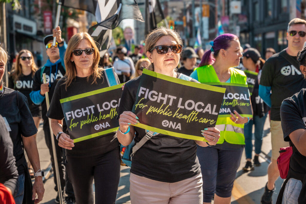 A crowd of people marches holding signs that say “Fight Local for public health care.”