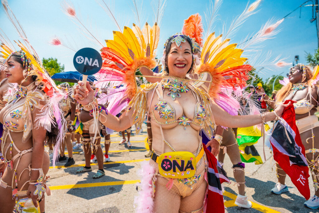 A ONA member in a Carnival costume holds an ONA fan and poses for the camera.