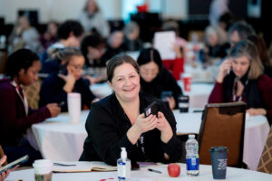 Woman sitting at table holding phone smiles at camera.