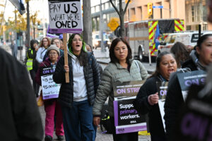 Members walk on picket line carrying and wearing signs.