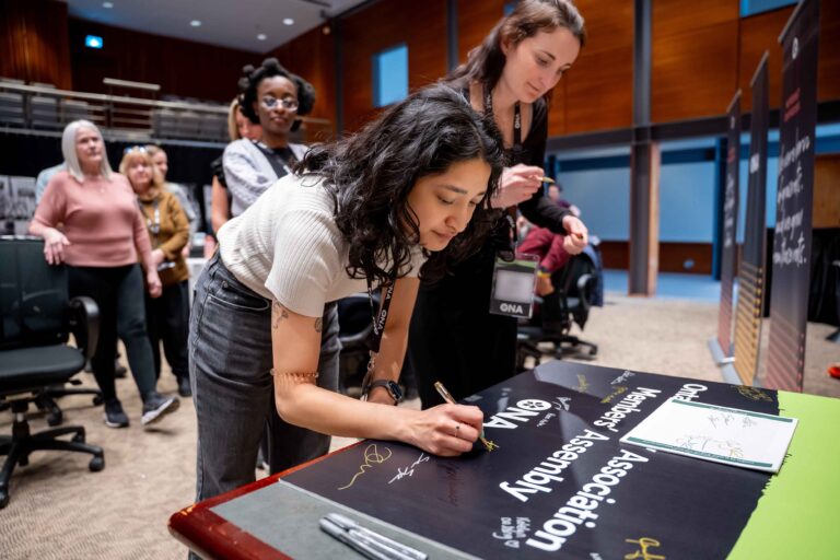 Members write their names on a Members’ Assembly sign.