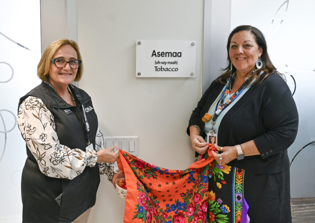 Two women stand in front of a plaque reading, “Asemma (Tobacco).”