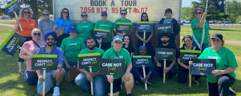 Group of members stand and kneel holding signs in front of a nursing home sign.