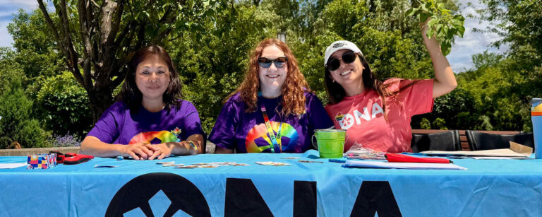 ONA6 Coordinator Jane Penciner sits between two fellow members at a table outdoors.