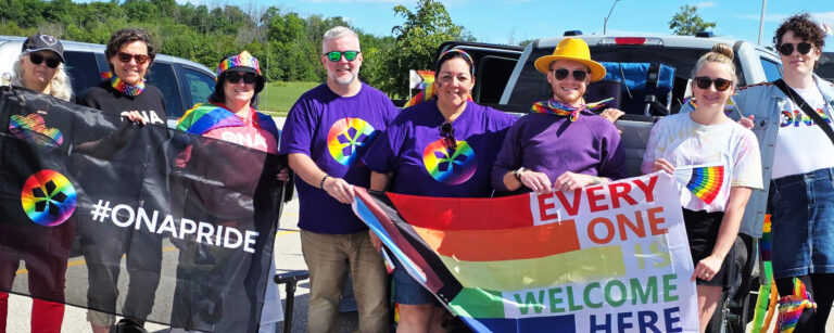 A group of ONA members, including Dustin Taylor-Brown, stand in a row wearing purple Pride t-shirts and holding Pride flags.