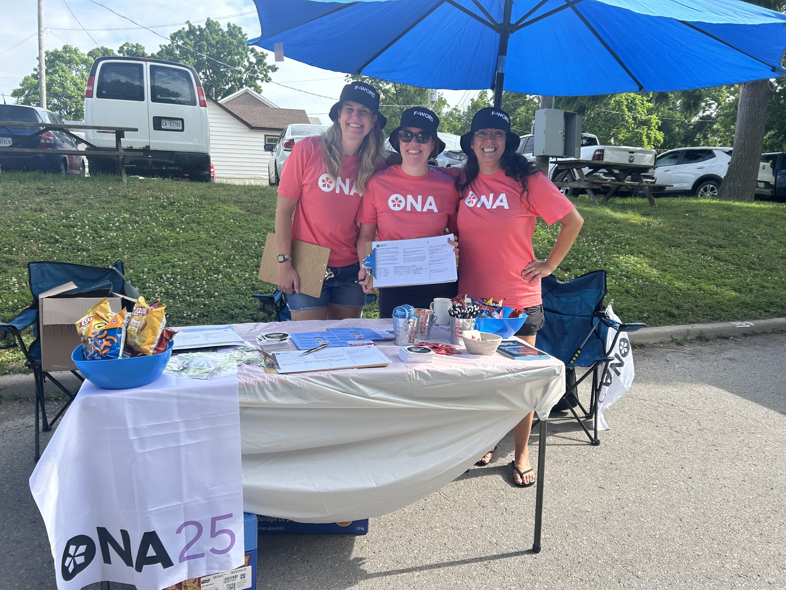 ONA25 members Paige Winton, Katherine Hattle and Liz Lima wear ONA t-shirts and F-Word bucket hats while holding ONA petitions at an outdoor table.