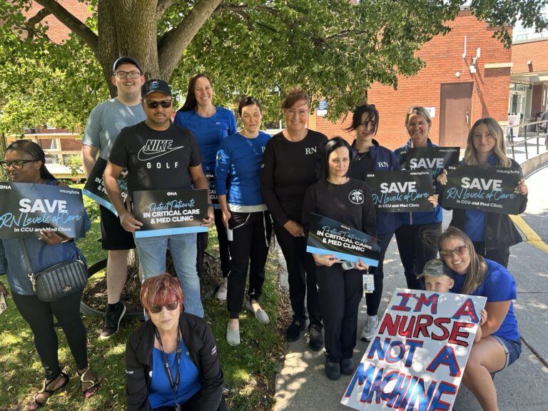 Group of members from Brant Community Healthcare System, wearing black and blue clothes and holding signs, stand under a tree.