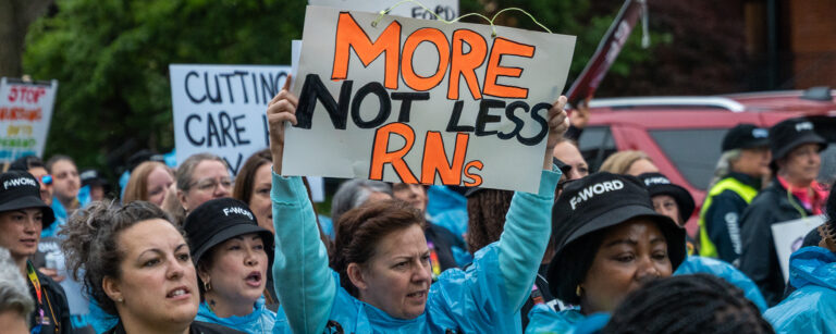 Members stand at a rally, with one holding up a sign that reads, “More, not Less RNs.”