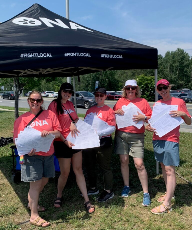 Group of members wearing ONA t-shirts stand in front of an outdoor table holding petitions.