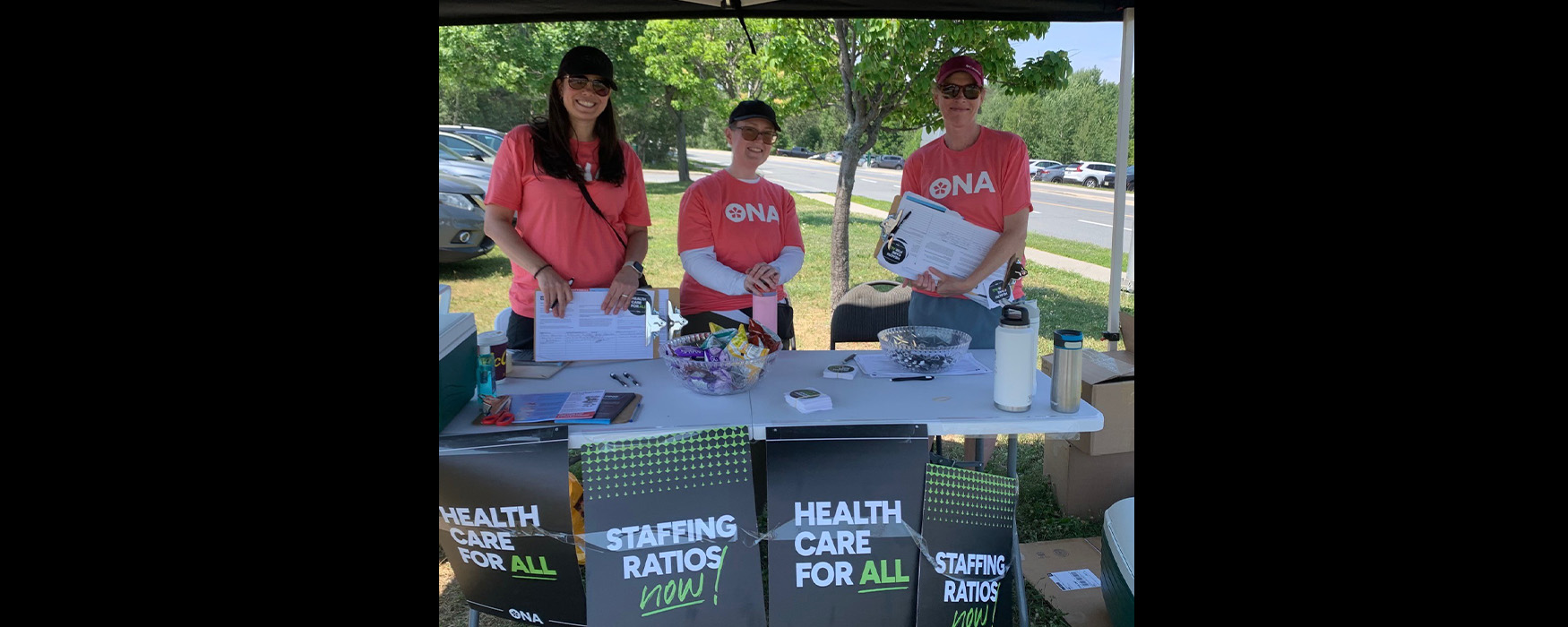 Three smiling members stand behind an ONA table displaying petitions, posters, snacks and stickers.